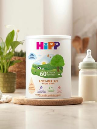 HiPP Organic Anti Reflux milk tub on a bright kitchen worktop beside a prepared baby bottle, dummy and soft toy