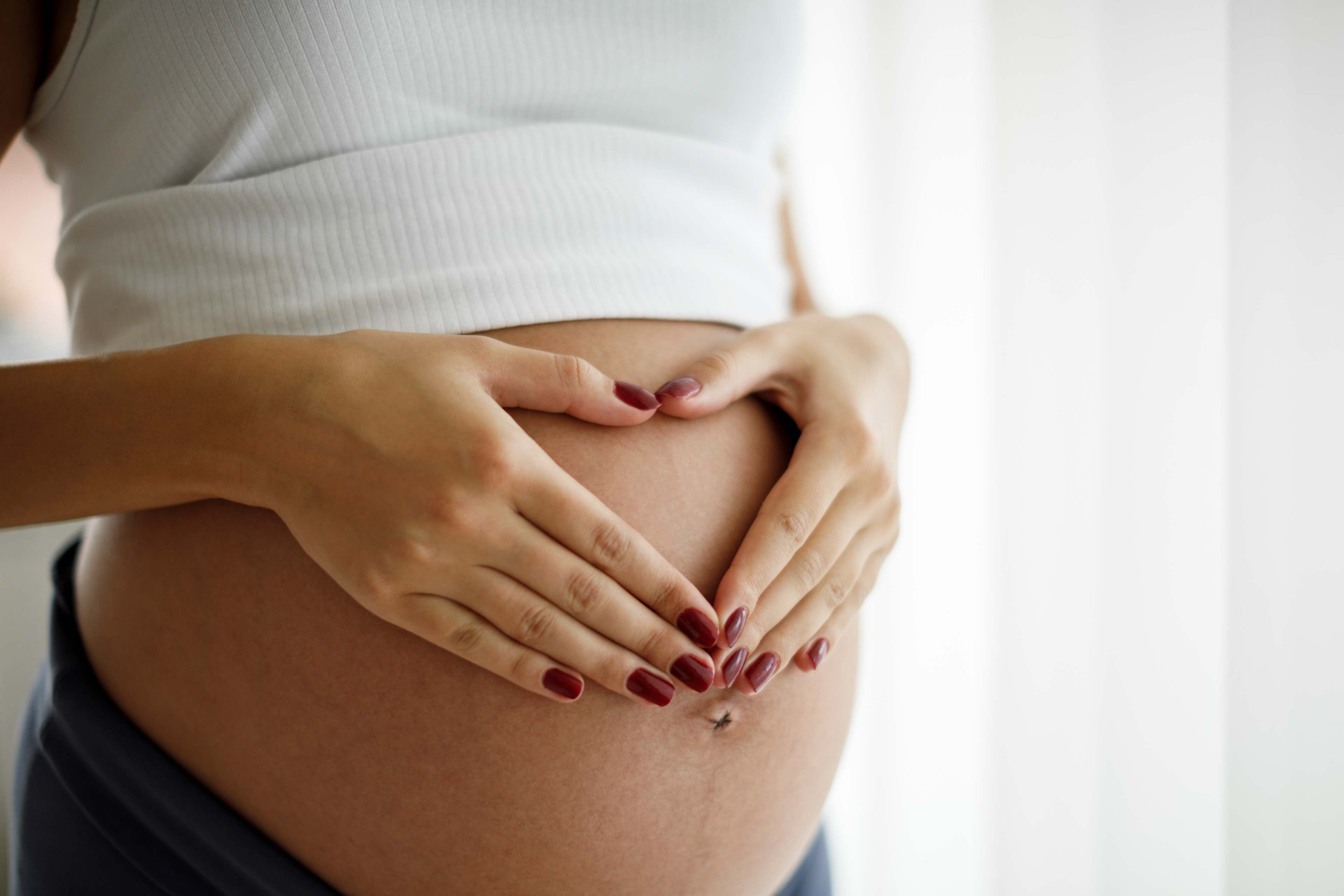 Schwangere Frau legt Hände mit roten Fingernägeln in Herzform auf ihren nackten Bauch