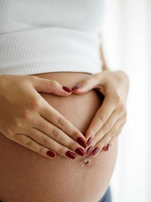 Schwangere Frau legt Hände mit roten Fingernägeln in Herzform auf ihren nackten Bauch