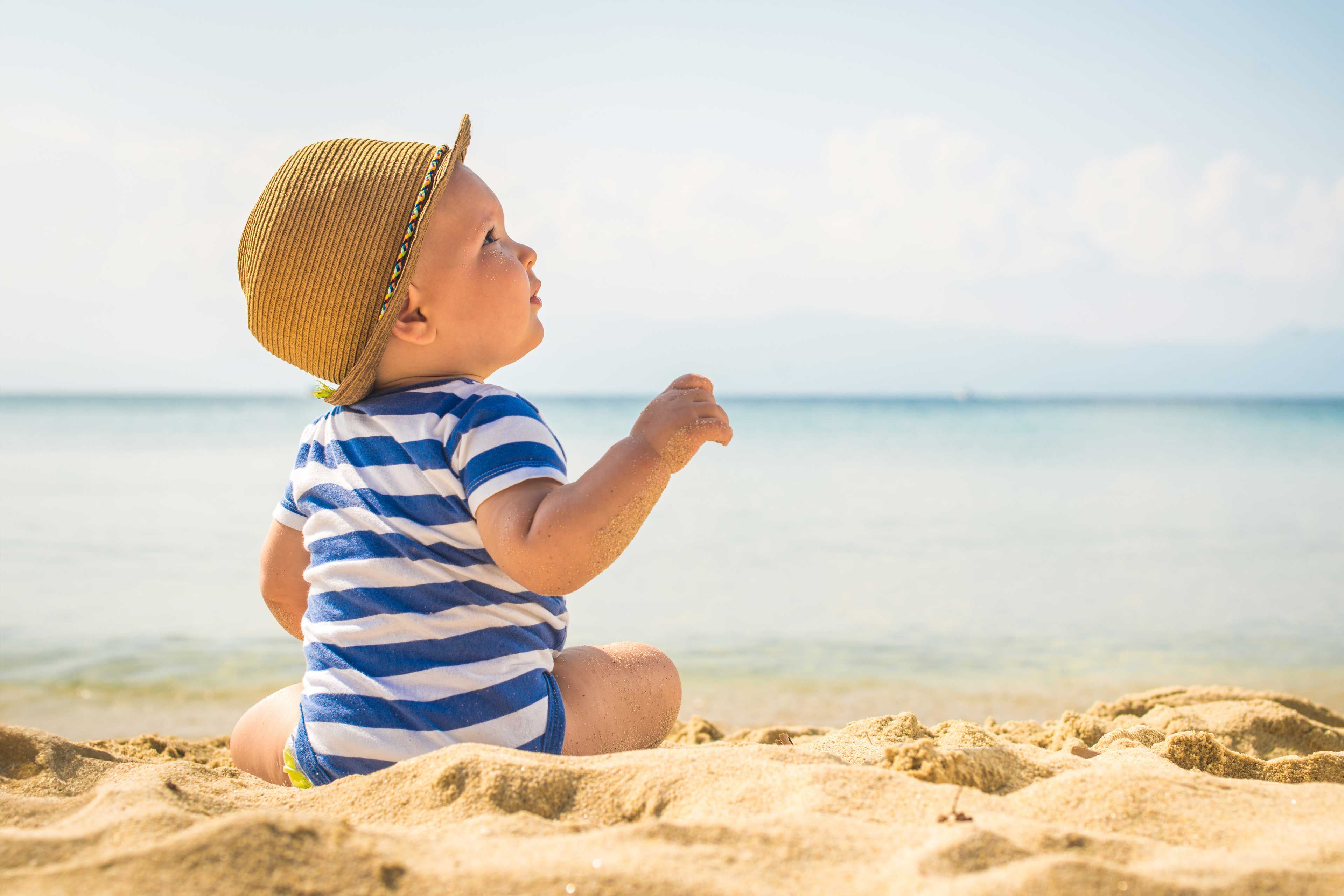 Kleinkind mit gestreiftem T-Shirt und Strohhut sitzt im Sand und blickt aufs Meer