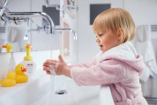 Ein kleines Kind in einem rosafarbenen Bademantel hält eine gelbe Gummiente unter einen Wasserhahn in einem Badezimmer.