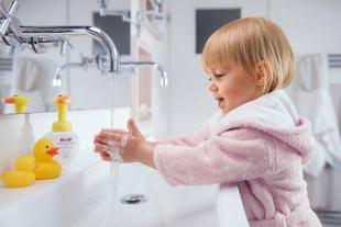 Ein kleines Kind in einem rosafarbenen Bademantel hält eine gelbe Gummiente unter einen Wasserhahn in einem Badezimmer.