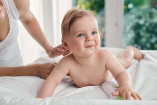 Ein Baby, das auf einer weißen Fläche liegt und neugierig nach oben schaut, während die Hand einer Person sanft seinen Rücken berührt.
