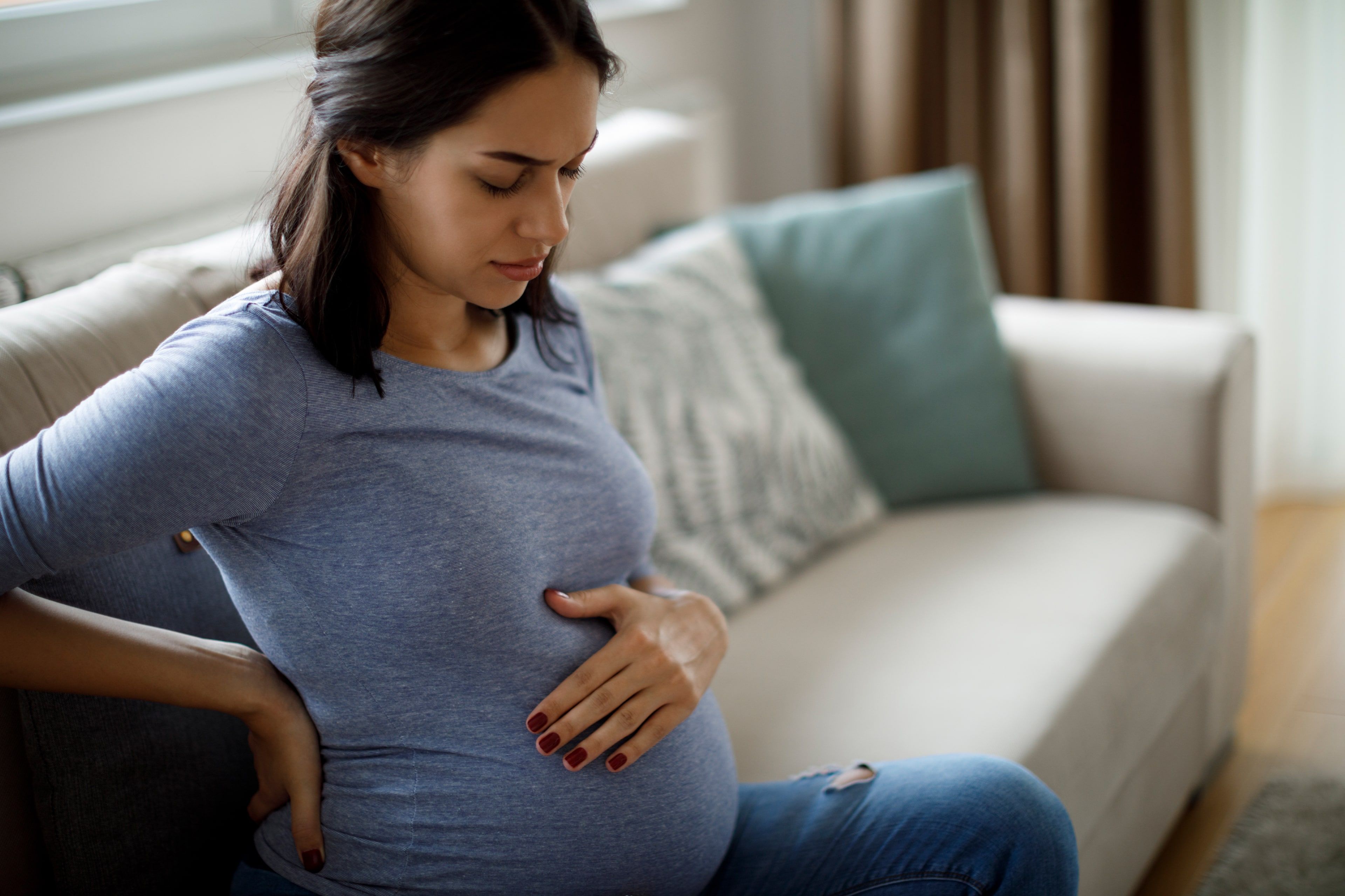 Eine Frau in einem blauen Oberteil, die sanft ihren Bauch berührt, mit einem nachdenklichen Gesichtsausdruck, sitzt auf einer Couch in einem Raum mit Fenster.