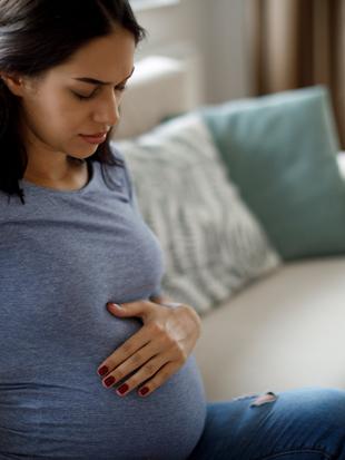 Eine Frau in einem blauen Oberteil, die sanft ihren Bauch berührt, mit einem nachdenklichen Gesichtsausdruck, sitzt auf einer Couch in einem Raum mit Fenster.