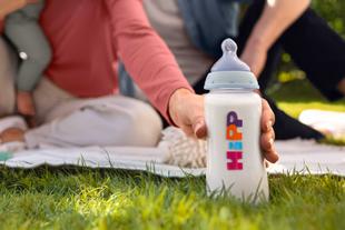 Babyflasche mit der Aufschrift 'HiPP' steht auf Gras, im Hintergrund unscharf sitzende Personen