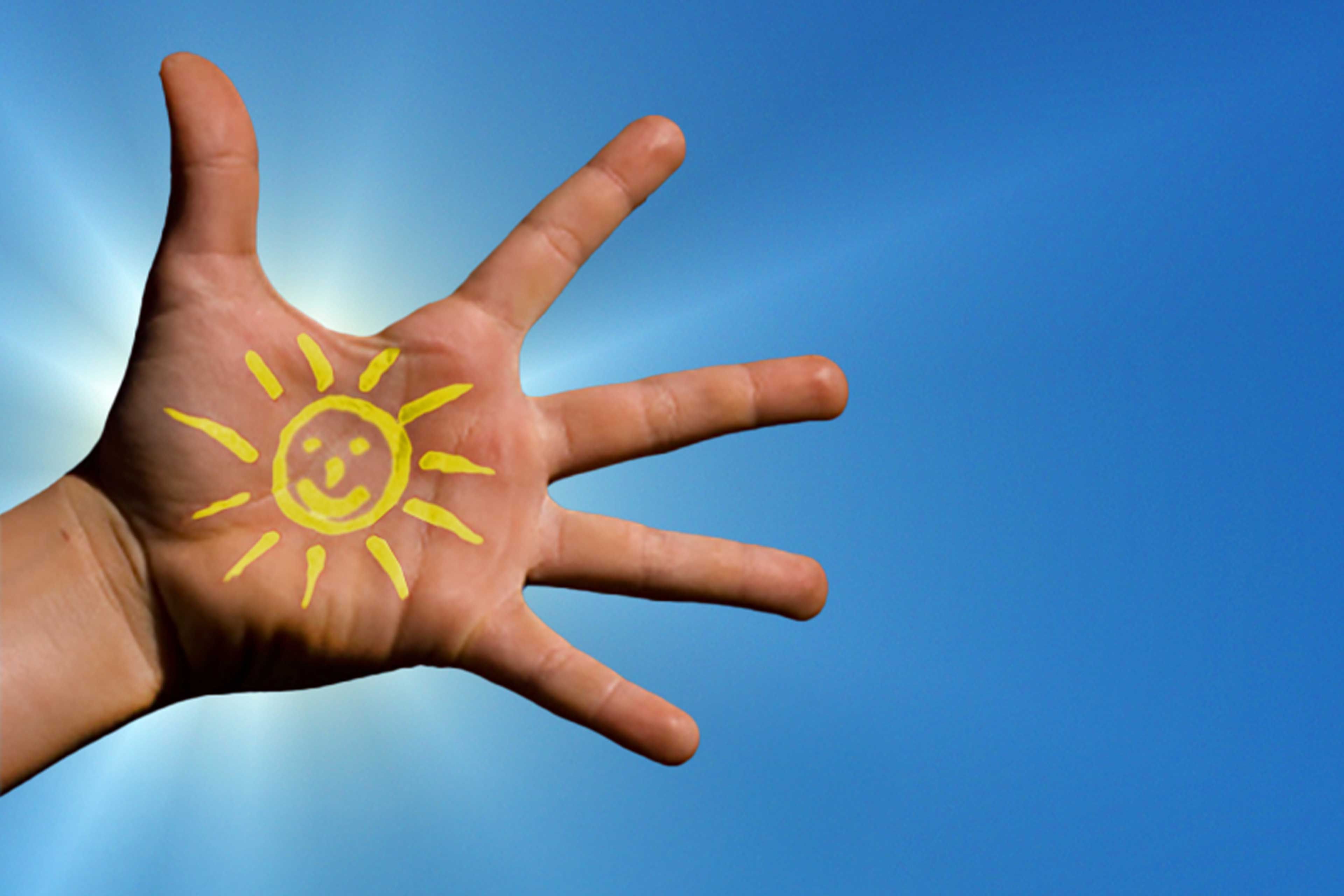 Geöffnete Hand mit gelber, lachender Sonne auf der Handfläche vor strahlend blauem Himmel als Symbol für Vitamin D