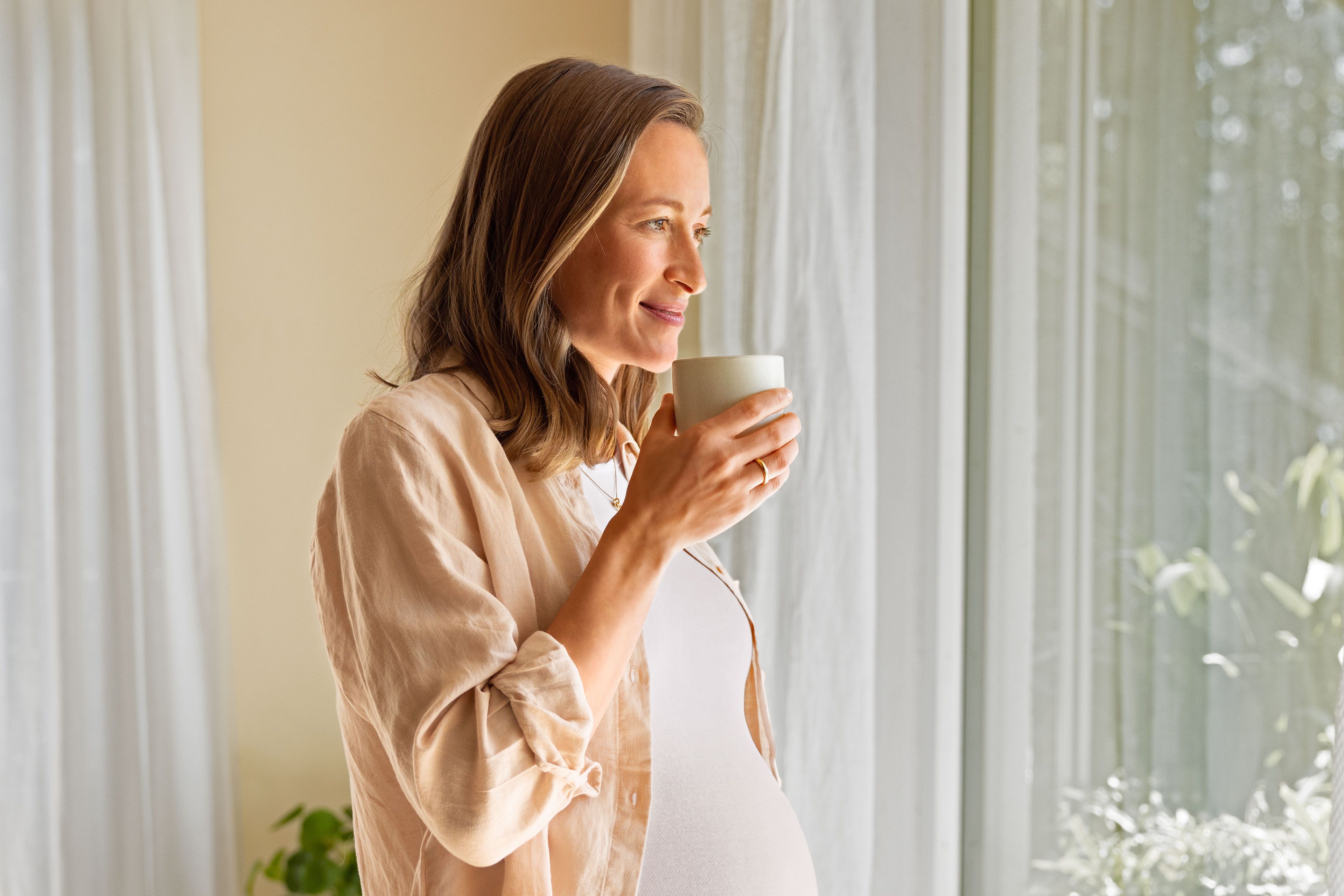 Eine Frau in einem beigen Hemd hält eine weiße Tasse und schaut aus einem Fenster mit durchsichtigen Vorhängen.