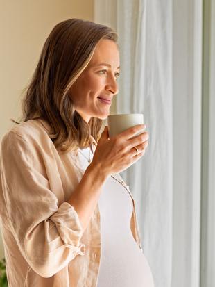 Eine Frau in einem beigen Hemd hält eine weiße Tasse und schaut aus einem Fenster mit durchsichtigen Vorhängen.