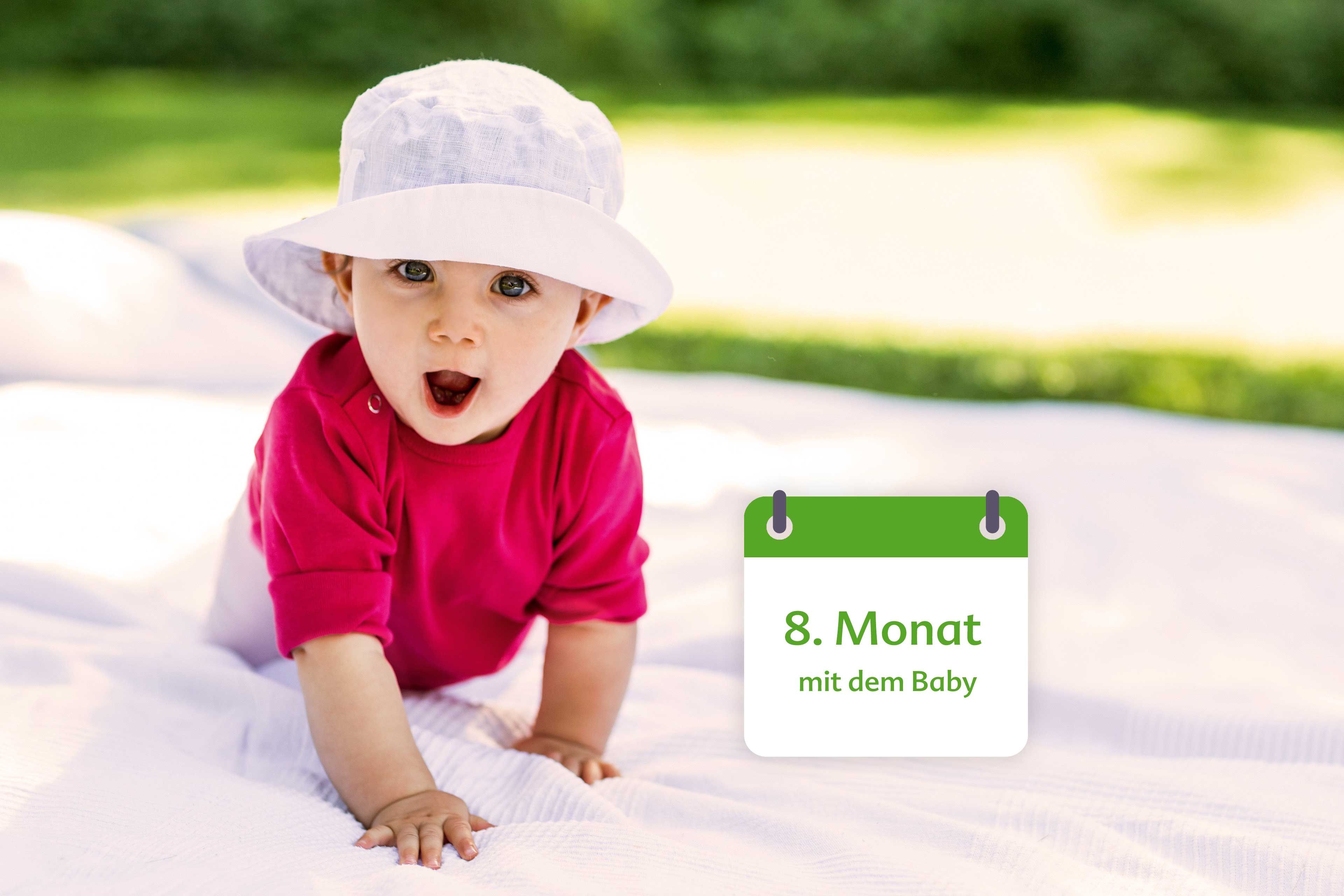 Ein Baby, bekleidet mit einem rosafarbenen Kleid und einem weißen Hut, liegt auf einer Decke im Freien und hat ein großes Kissen neben sich.