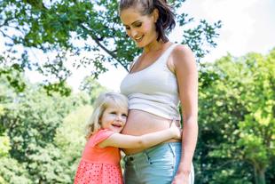 Schwangere Mutter im Park, deren kleines Kind liebevoll den Babybauch umarmt und sich auf das Geschwisterchen freut