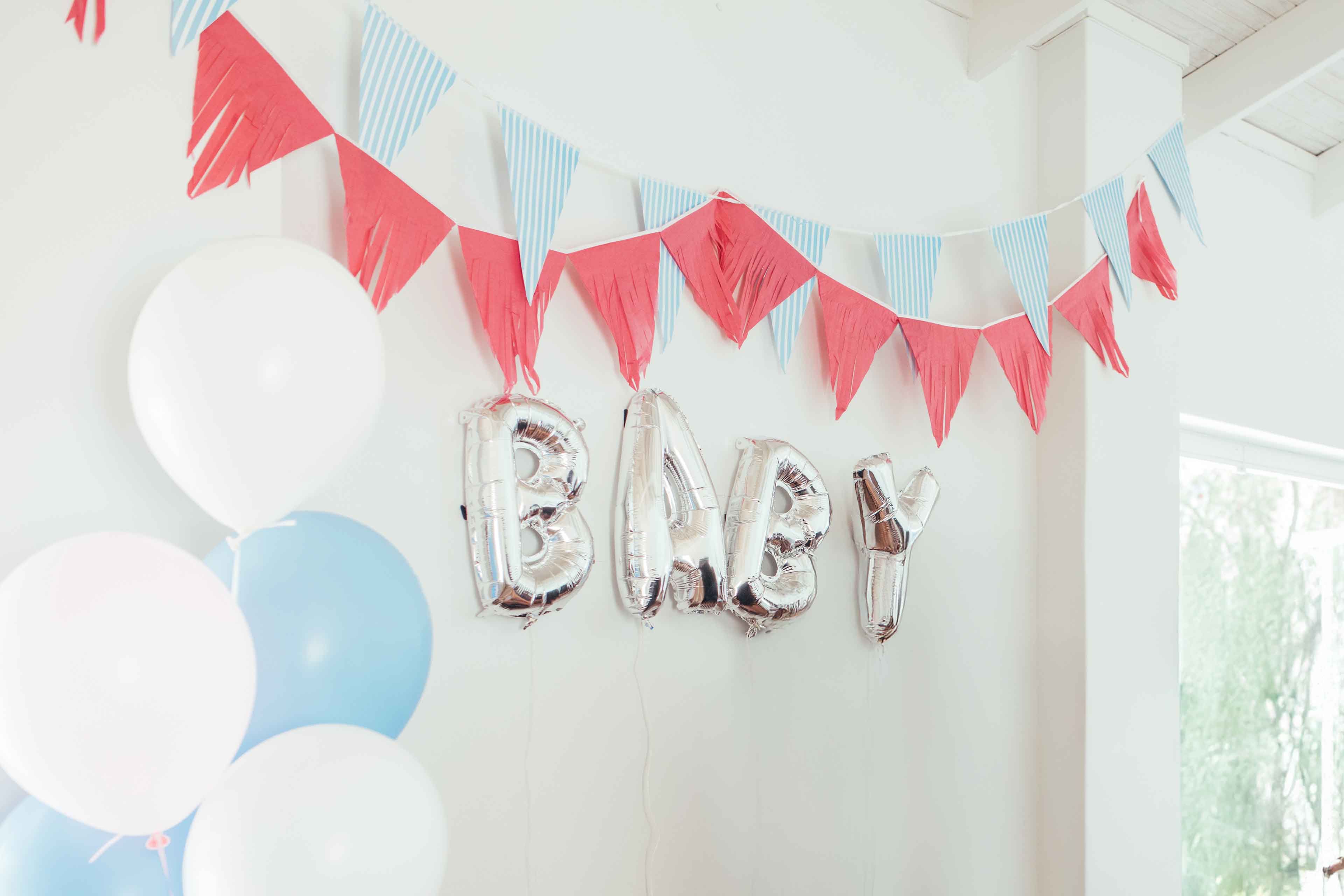 Silberne Luftballonbuchstaben bilden das Wort BABY an einer Wand mit roten und blau-weißen Wimpelgirlanden und weißen sowie blauen Luftballons