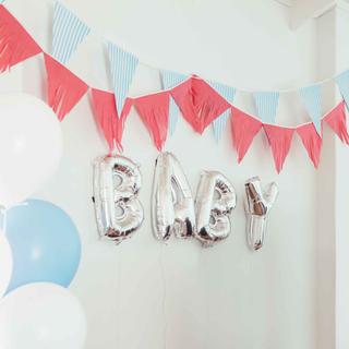 Silberne Luftballonbuchstaben bilden das Wort BABY an einer Wand mit roten und blau-weißen Wimpelgirlanden und weißen sowie blauen Luftballons