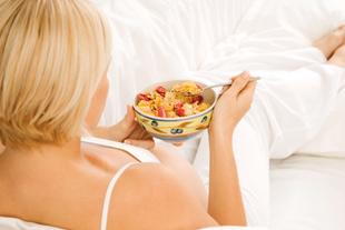 Eine Frau mit blondem Haar, in weißer Kleidung, hält eine Schüssel mit Müsli und verschiedenen Früchten in der Hand und liegt auf einer weißen Fläche.