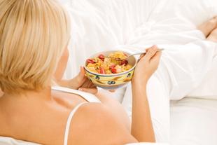 Eine Frau mit blondem Haar, in weißer Kleidung, hält eine Schüssel mit Müsli und verschiedenen Früchten in der Hand und liegt auf einer weißen Fläche.