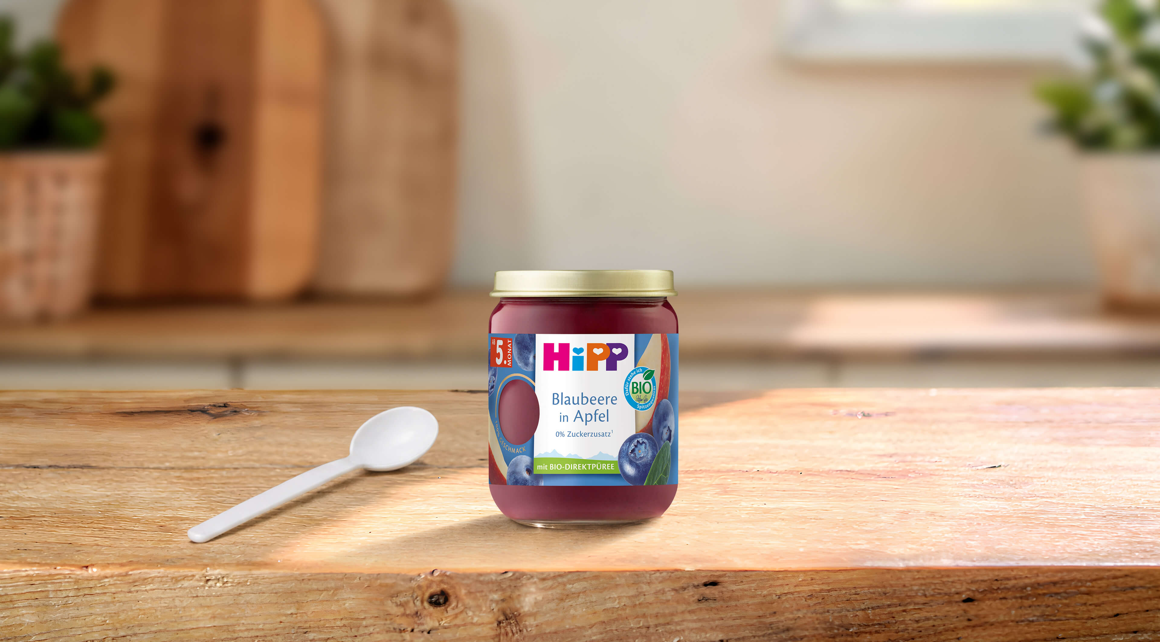 HiPP Bio-Gläschen „Blaubeere in Apfel“ ab 5. Monat auf Holztisch mit Löffel in heller, gemütlicher Küchenumgebung