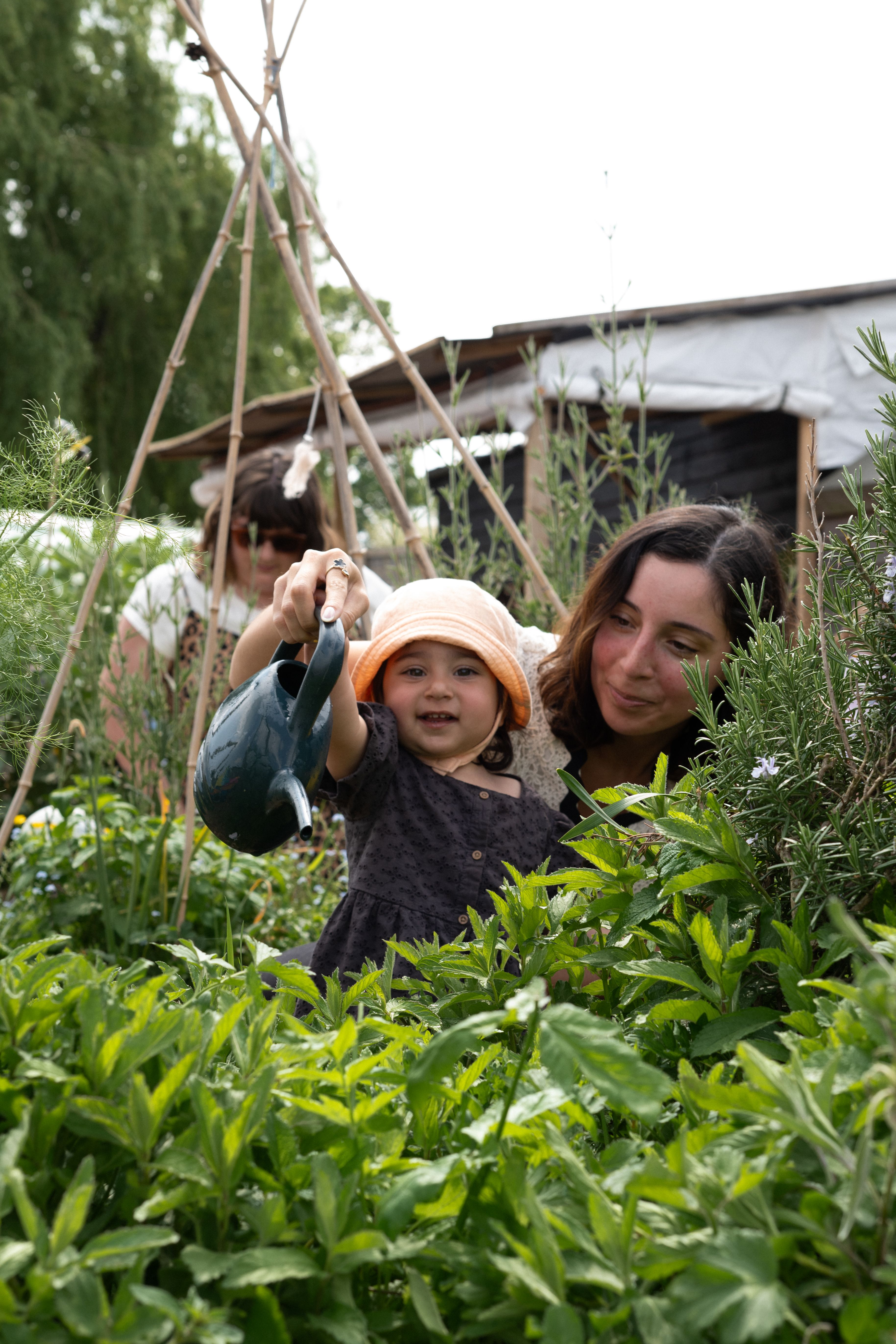 Smiling toddler watering plants in a lush outdoor garden with a parent beside them