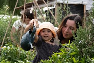 Smiling toddler watering plants in a lush outdoor garden with a parent beside them