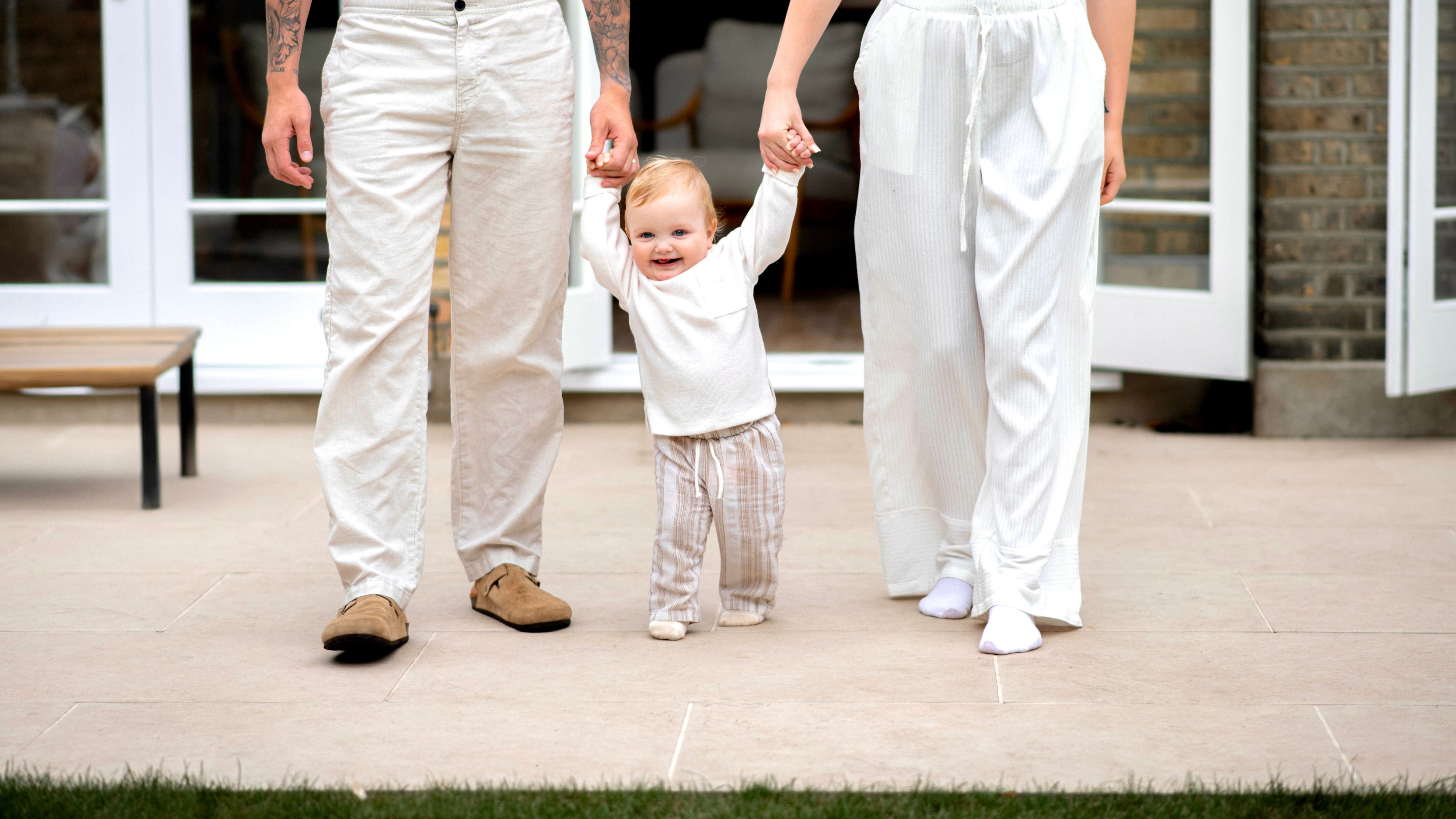 Smiling toddler learning to walk outside, holding hands with two parents in light, relaxed clothing.