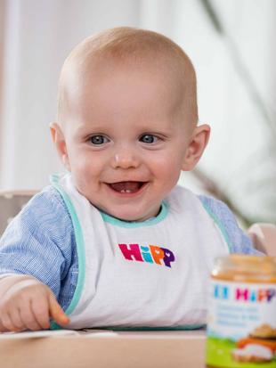 Ein Baby in einem Hochstuhl, das ein Lätzchen mit der Aufschrift "HIPP" trägt und daneben ein Glas mit Babynahrung.