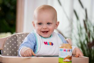 Lachendes Baby im Hochstuhl mit HiPP-Lätzchen und einem Glas Babynahrung vor sich