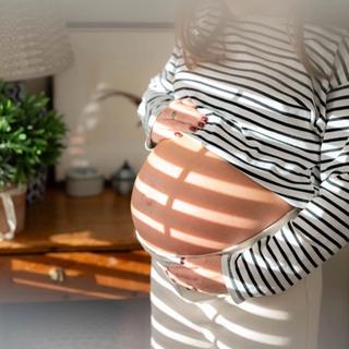 A close-up of a pregnant woman holding her bare belly as sunlight filters through window blinds, casting striped shadows across her skin. She’s wearing a black-and-white striped top and cream pants in a softly lit room.