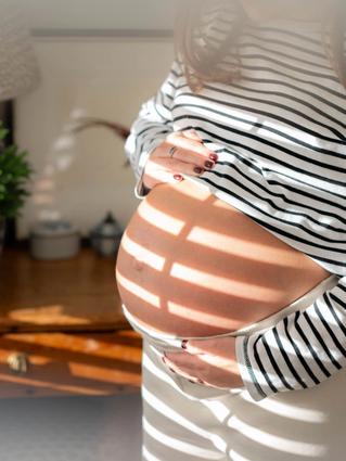 A close-up of a pregnant woman holding her bare belly as sunlight filters through window blinds, casting striped shadows across her skin. She’s wearing a black-and-white striped top and cream pants in a softly lit room.
