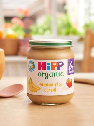 HiPP Organic Banana Rice Cereal jar on a dining table next to a bowl and spoon