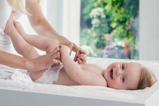 Ein fröhliches Baby, das auf einer weißen Fläche liegt und dessen Beine von den Händen einer Person sanft gehalten werden, vor einem hellen und luftigen Hintergrund.