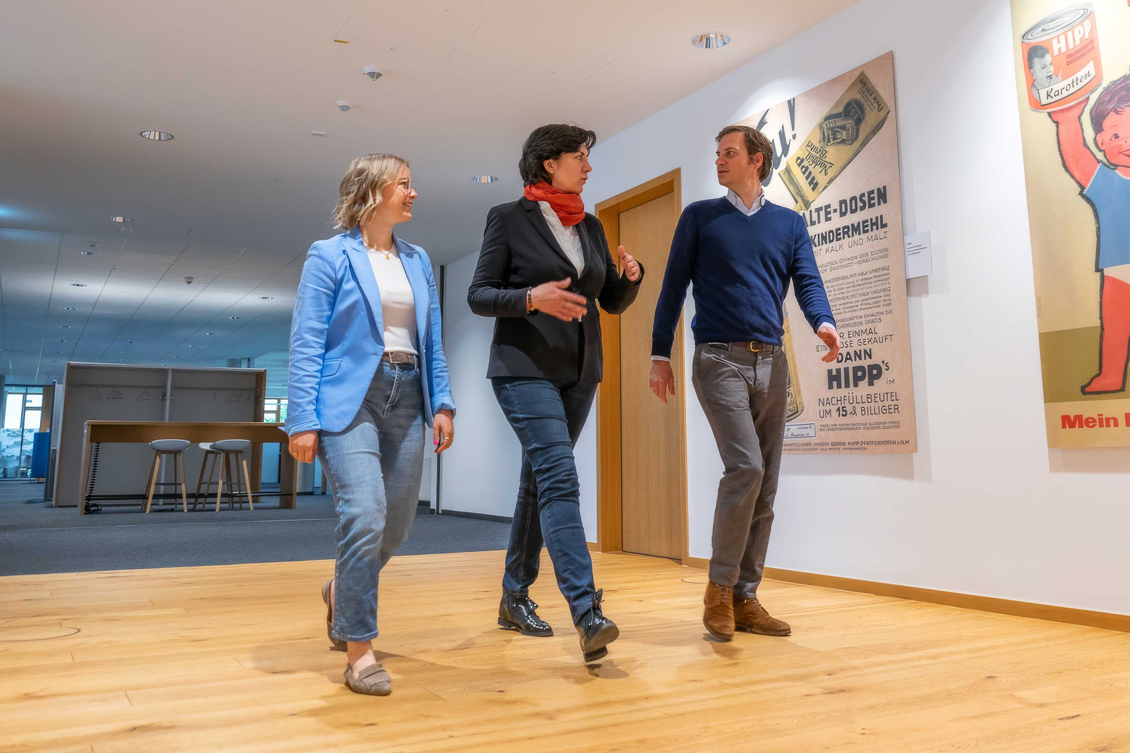 Drei HiPP Mitarbeitende gehen in einem hellen Flur an historischen Werbeplakaten vorbei