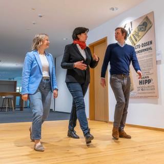 Drei HiPP Mitarbeitende gehen in einem hellen Flur an historischen Werbeplakaten vorbei