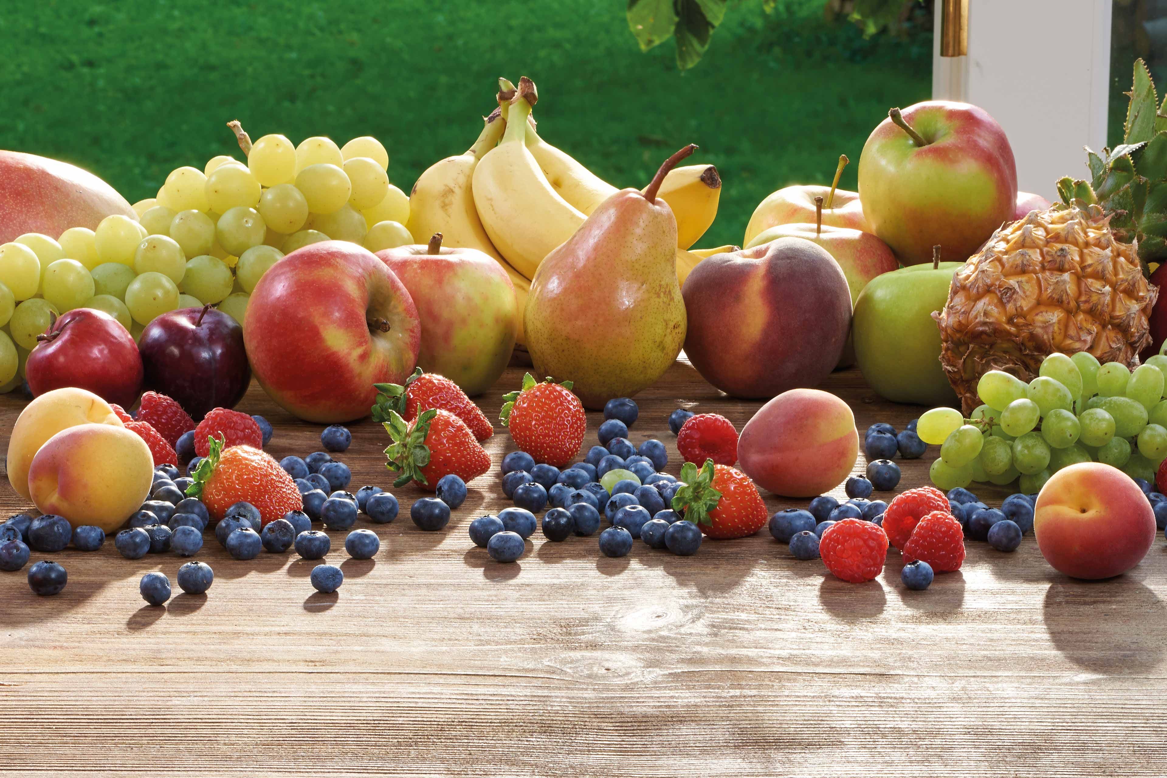 Eine Vielzahl von frischen Früchten und Beeren, die auf einer hölzernen Oberfläche angeordnet sind, darunter Äpfel, Bananen, Trauben, Birnen, Erdbeeren, Heidelbeeren und eine Ananas.