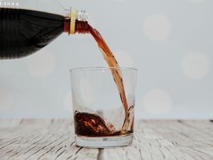 Ein klares Glas, das mit einer braunen Flüssigkeit gefüllt ist, bei der es sich vermutlich um Soda handelt, wird aus einer dunklen Flasche gegossen.