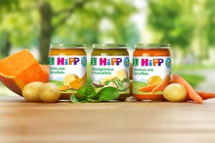 Drei Gläschen Babynahrung mit der Aufschrift 'Hipp' mit verschiedenen Gemüsekombinationen, begleitet von frischem Gemüse und Obst.