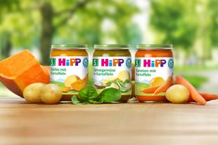 Drei Gläschen Babynahrung mit der Aufschrift 'Hipp' mit verschiedenen Gemüsekombinationen, begleitet von frischem Gemüse und Obst.