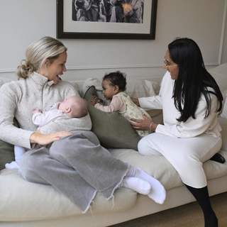 Two mothers sit on a cozy beige couch, engaging with their babies. One woman, wearing a light gray sweater, is breastfeeding her infant while smiling at her friend. The other woman, dressed in a white outfit, gently supports a baby in a floral onesie who is sitting upright on the couch. The room has a warm, inviting feel with soft cushions and a framed black-and-white artwork on the wall.