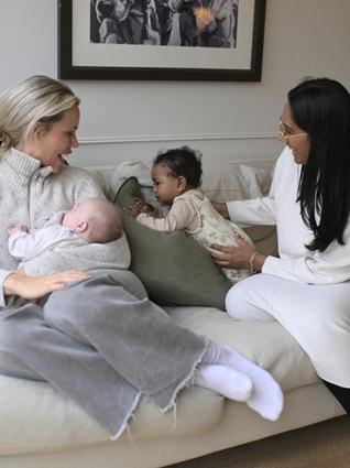 Two mothers sit on a cozy beige couch, engaging with their babies. One woman, wearing a light gray sweater, is breastfeeding her infant while smiling at her friend. The other woman, dressed in a white outfit, gently supports a baby in a floral onesie who is sitting upright on the couch. The room has a warm, inviting feel with soft cushions and a framed black-and-white artwork on the wall.