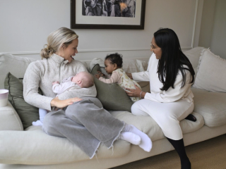 Two mothers sit on a cozy beige couch, engaging with their babies. One woman, wearing a light gray sweater, is breastfeeding her infant while smiling at her friend. The other woman, dressed in a white outfit, gently supports a baby in a floral onesie who is sitting upright on the couch. The room has a warm, inviting feel with soft cushions and a framed black-and-white artwork on the wall.