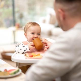 a 17 months old toddler holding a cup to its mouth and smiling to the camera with the father blurred in the front of the image.