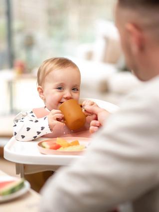 a 17 months old toddler holding a cup to its mouth and smiling to the camera with the father blurred in the front of the image.