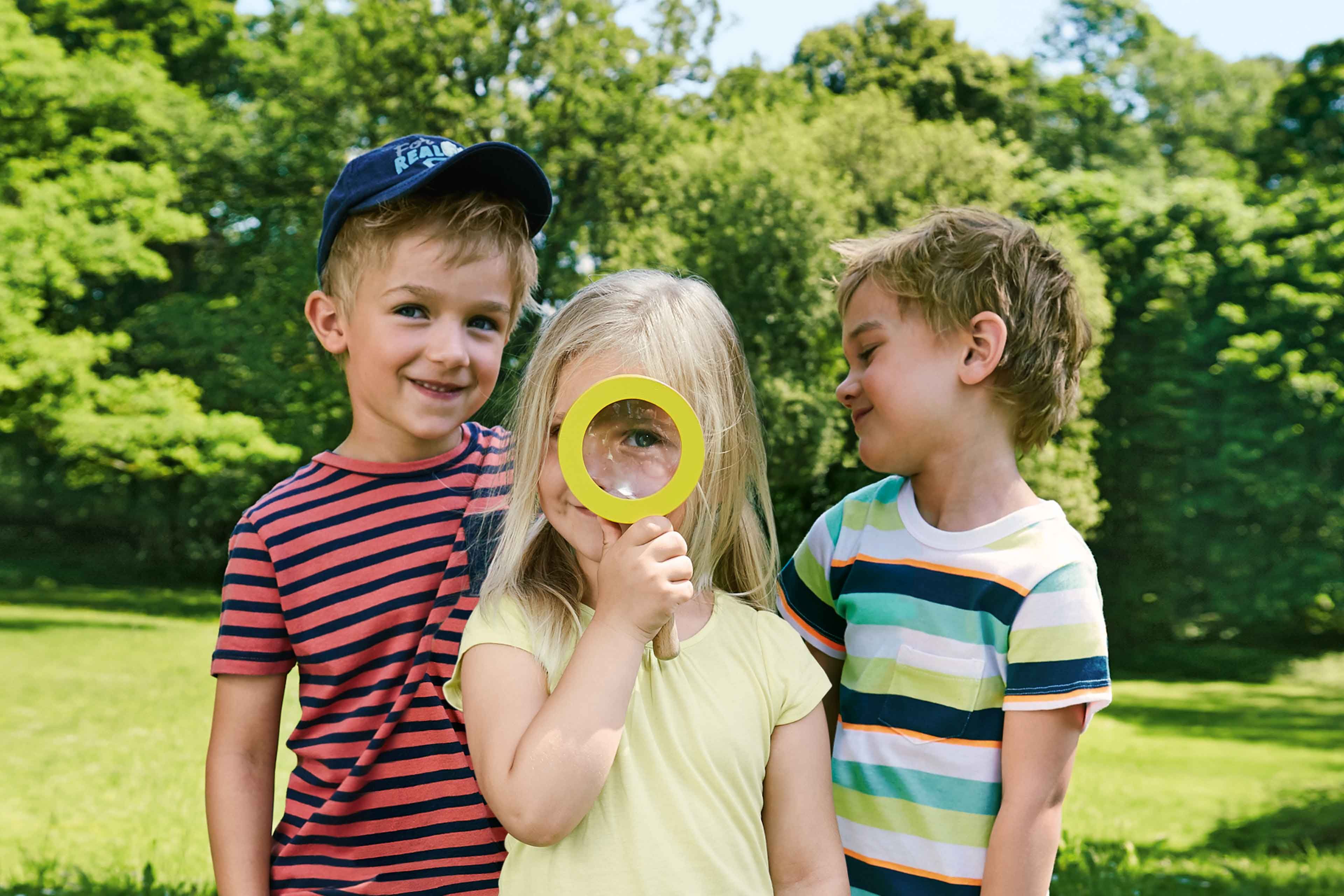 Drei Kinder im Freien, wobei ein Kind eine gelbe Lupe über sein Auge hält, während die beiden anderen es anschauen.