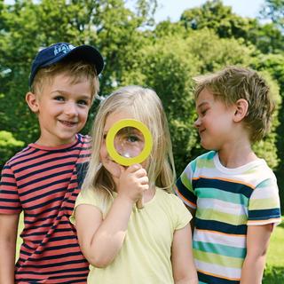 Drei Kinder im Freien, wobei ein Kind eine gelbe Lupe über sein Auge hält, während die beiden anderen es anschauen.