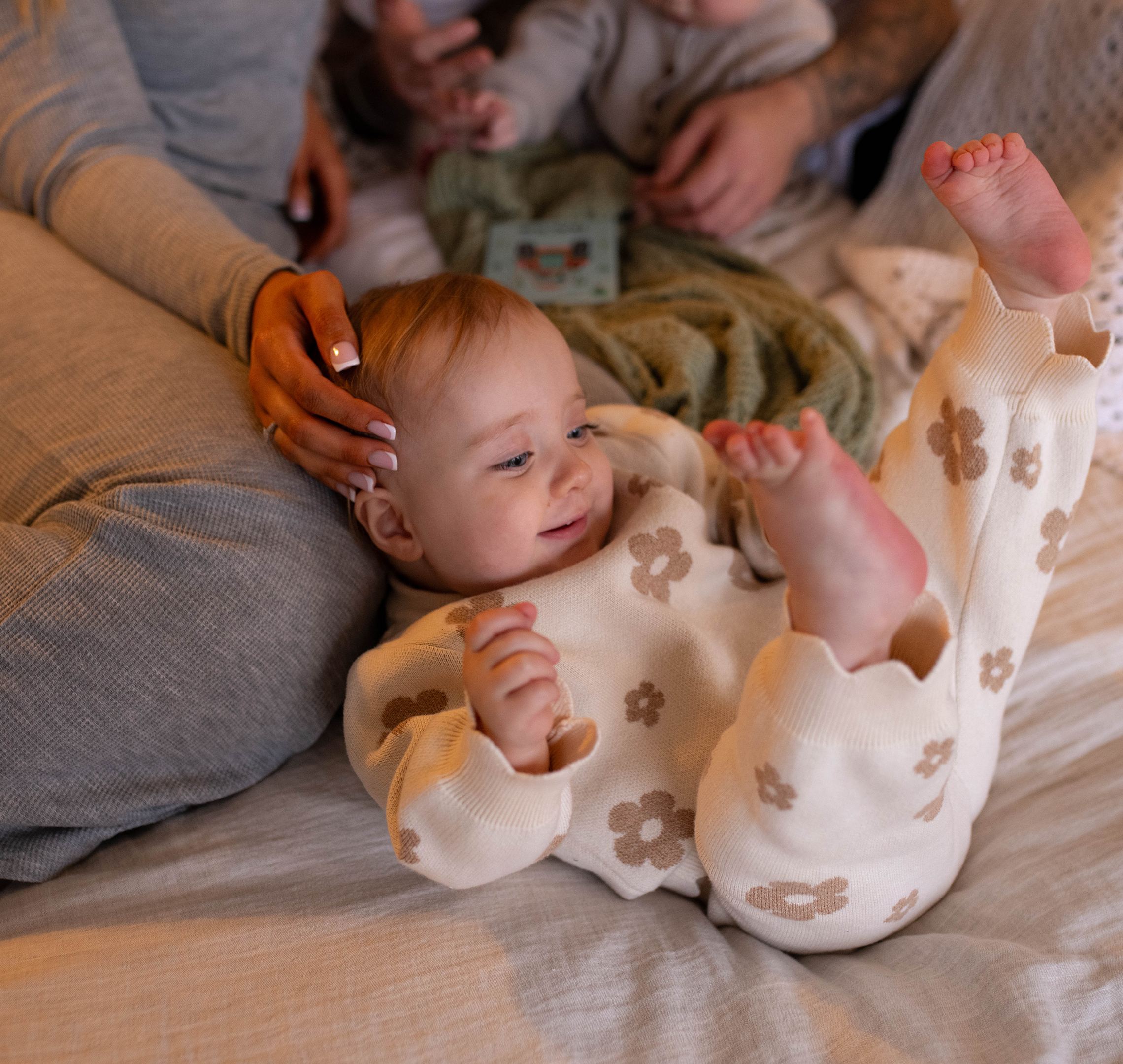 Smiling baby in floral pajamas lying on a bed, surrounded by family, conveying warmth and comfort.