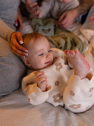 Smiling baby in floral pajamas lying on a bed, surrounded by family, conveying warmth and comfort.