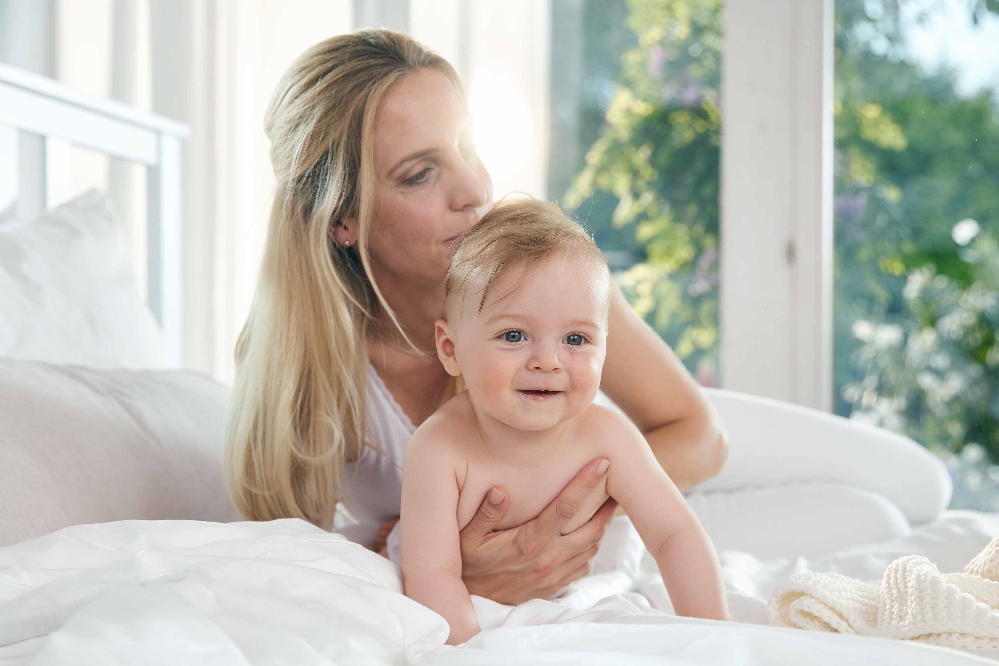 Eine Frau, die ein lächelndes Baby auf einem Bett hält, mit Blick durch das Fenster ins Freie.