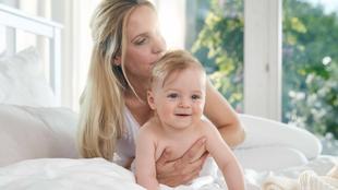 Eine Frau, die ein lächelndes Baby auf einem Bett hält, mit Blick durch das Fenster ins Freie.