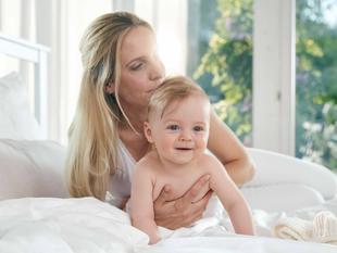 Eine Frau, die ein lächelndes Baby auf einem Bett hält, mit Blick durch das Fenster ins Freie.