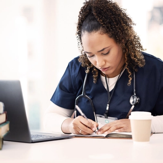Healthcare professional studying at a desk, writing notes beside a laptop as part of continued professional development