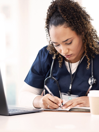 Healthcare professional studying at a desk, writing notes beside a laptop as part of continued professional development