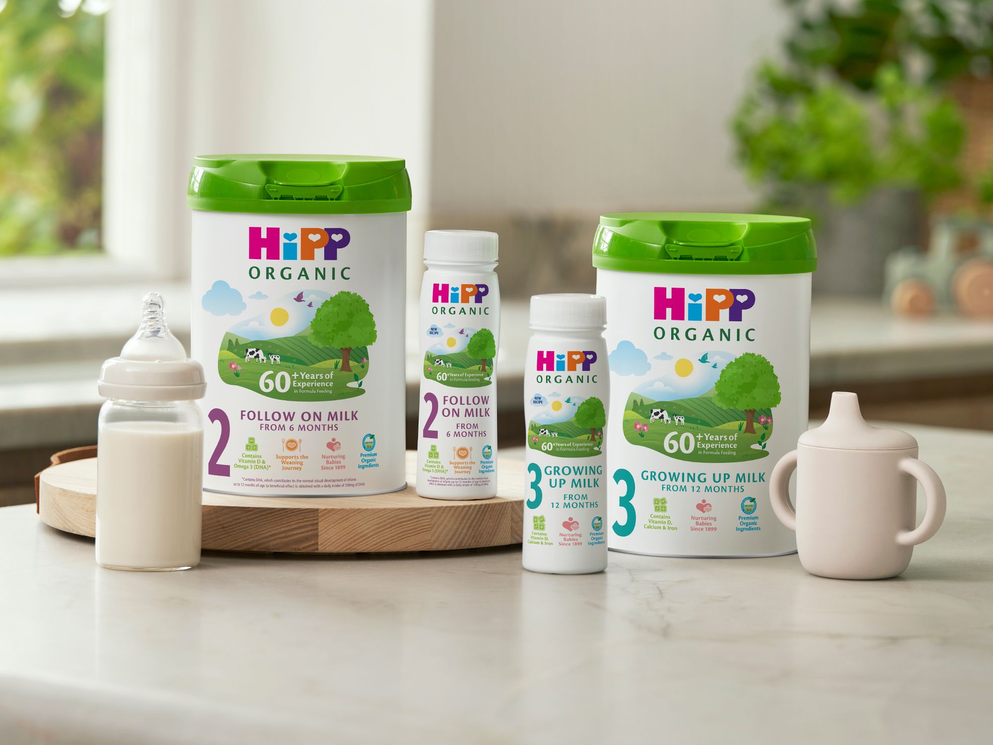 HiPP Organic follow on and growing up milks on a bright kitchen worktop beside a baby bottle and pastel sippy cup